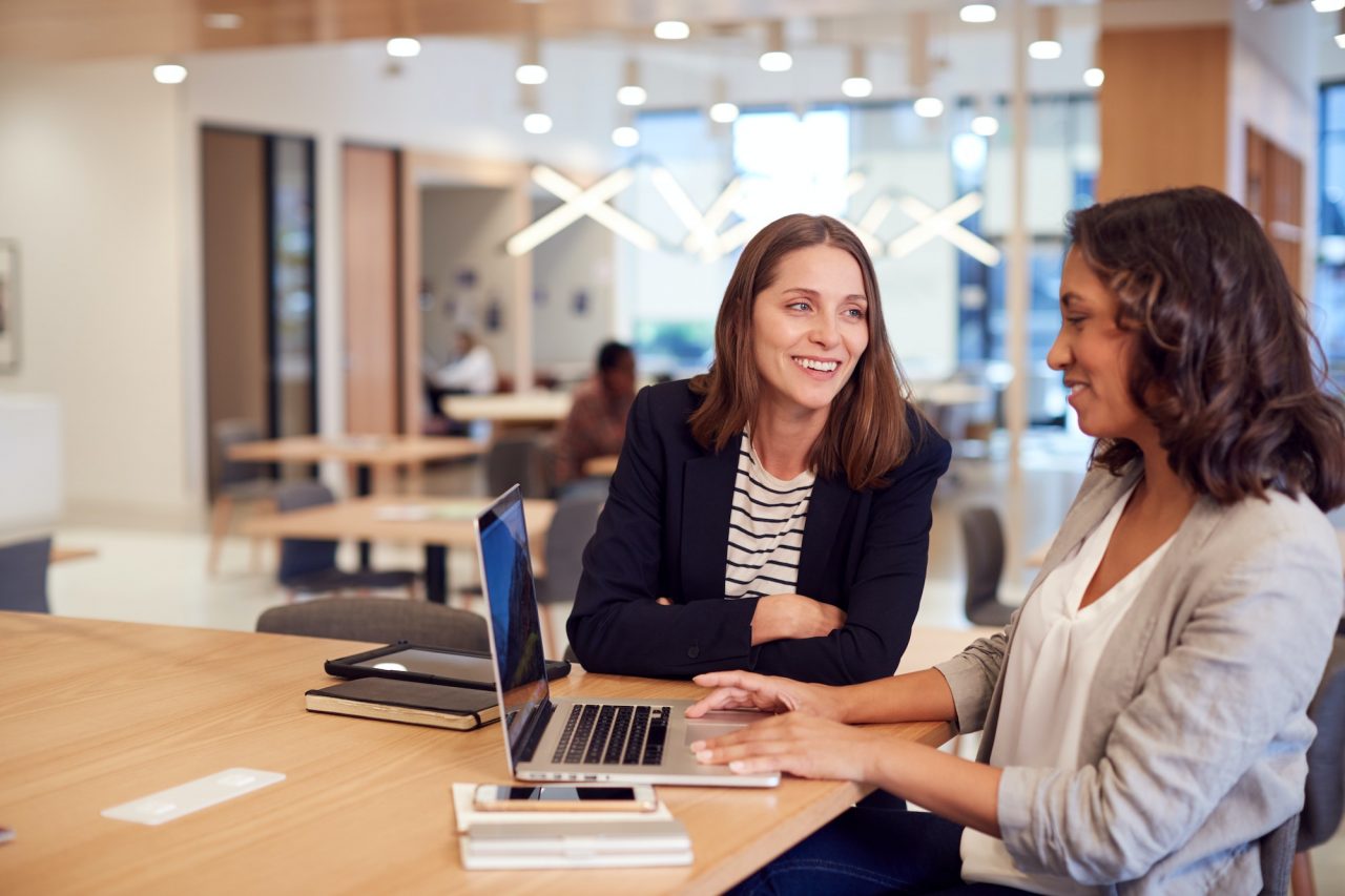 two businesswomen with laptop at desk in open plan office collaborating on project together